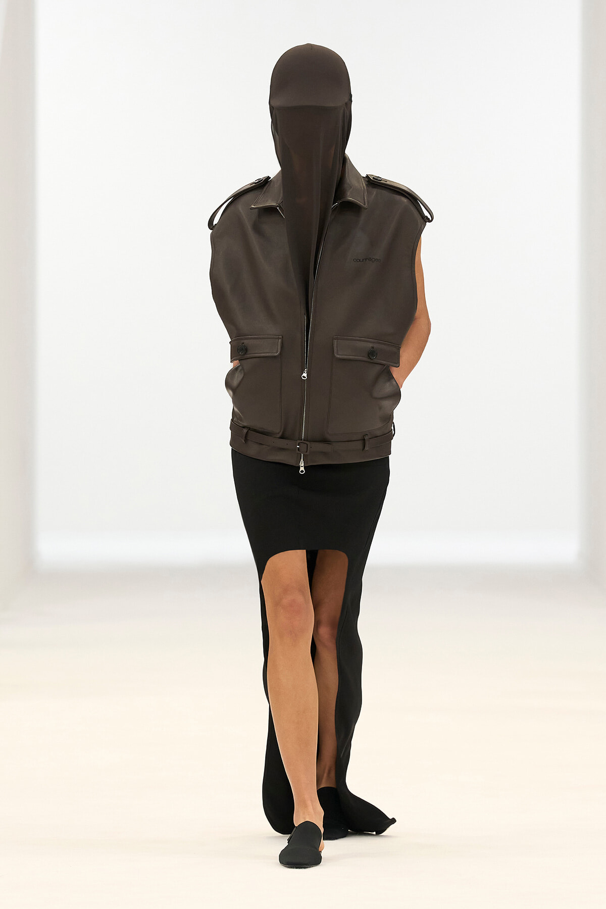 Model walking the runway in a dark brown sleeveless leather vest with a full-face veil and matching belt, paired with a black high-slit pencil skirt and black flats.