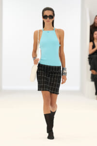 Model on a runway wearing a turquoise halter top, black grid-pattern skirt, black knee-high boots, dark sunglasses, and a white shoulder bag.
