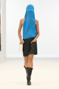 Model walking away in a bright blue sleeveless top and matching head veil, black leather mini skirt, knee-high boots, and a white shoulder bag.