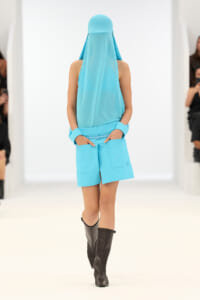 Runway model wearing a bright turquoise sleeveless top and matching shorts with pockets, paired with black knee-high boots.
