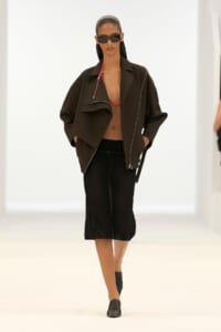 Model on a white runway wearing an oversized black jacket, red bralette, black culottes, black loafers, and sunglasses.