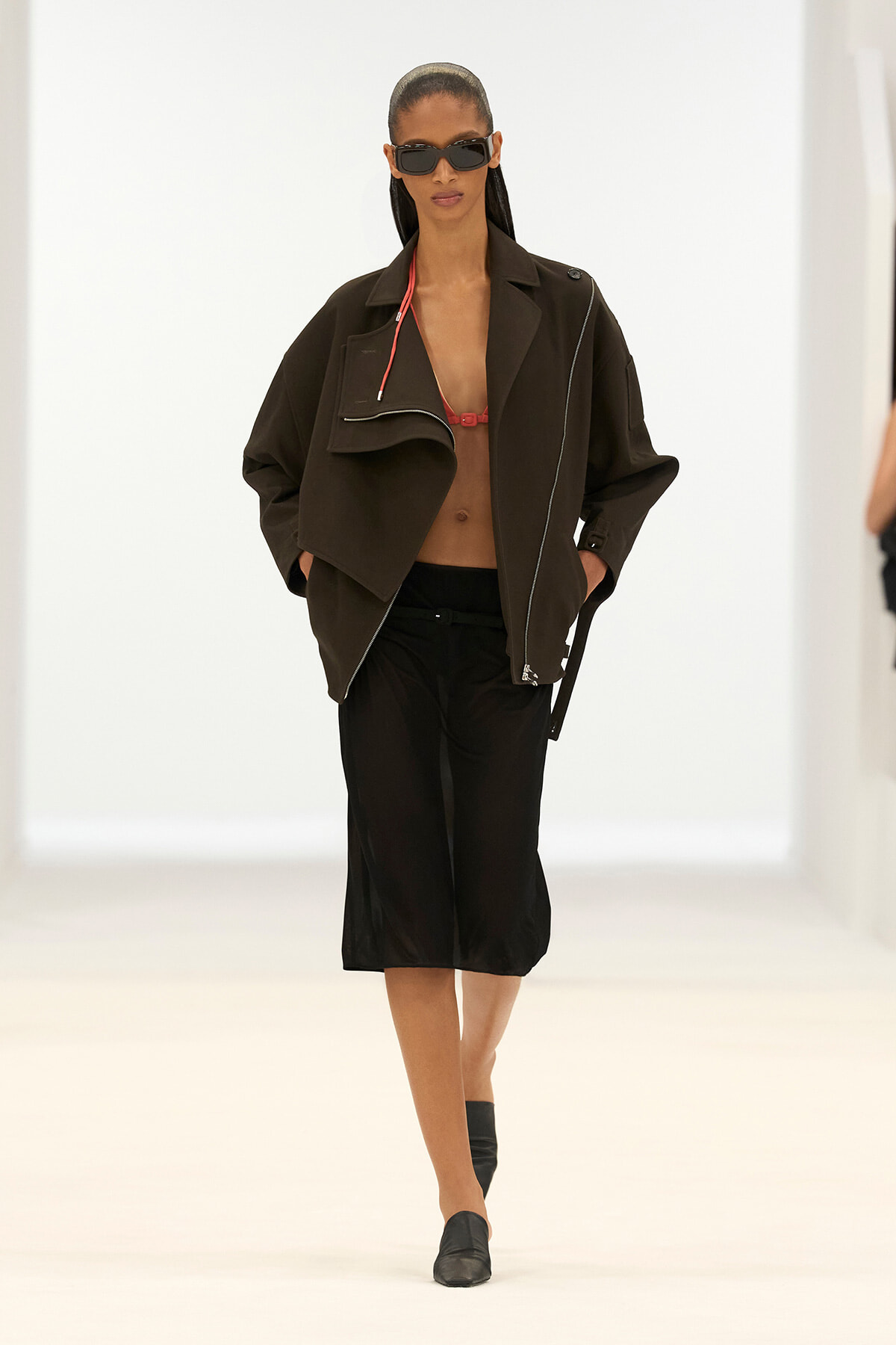 Model on a white runway wearing an oversized black jacket, red bralette, black culottes, black loafers, and sunglasses.