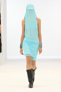 Model walking a fashion show wearing a turquoise sleeveless dress with a sheer headscarf and matching skirt, paired with silver cuffs and black knee-high boots from the back view.