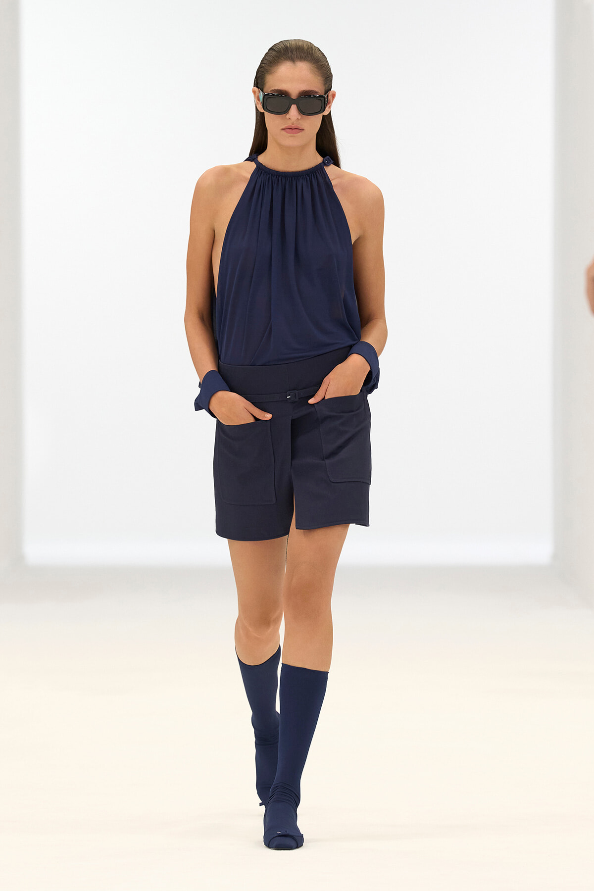 Model walking a runway in a navy halter top, matching shorts, navy knee-high socks, and sunglasses.