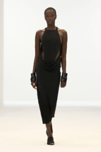 Model walking a minimalist black halter dress with draped sides and cutouts, on a white runway.