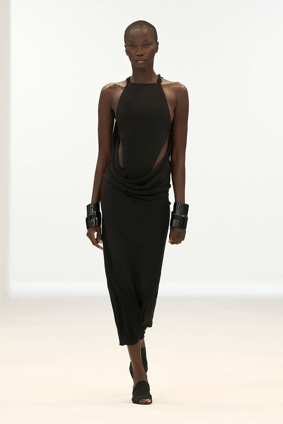 Model walking a minimalist black halter dress with draped sides and cutouts, on a white runway.