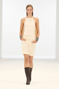 Model walking the runway in a sleeveless cream halter dress with a slim belt and knee-high black boots, wearing chunky silver cuffs.