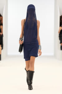 Model walking runway in a navy blue sleeveless dress with sheer veil and cap, paired with tall black boots and chunky silver cuffs on wrists.