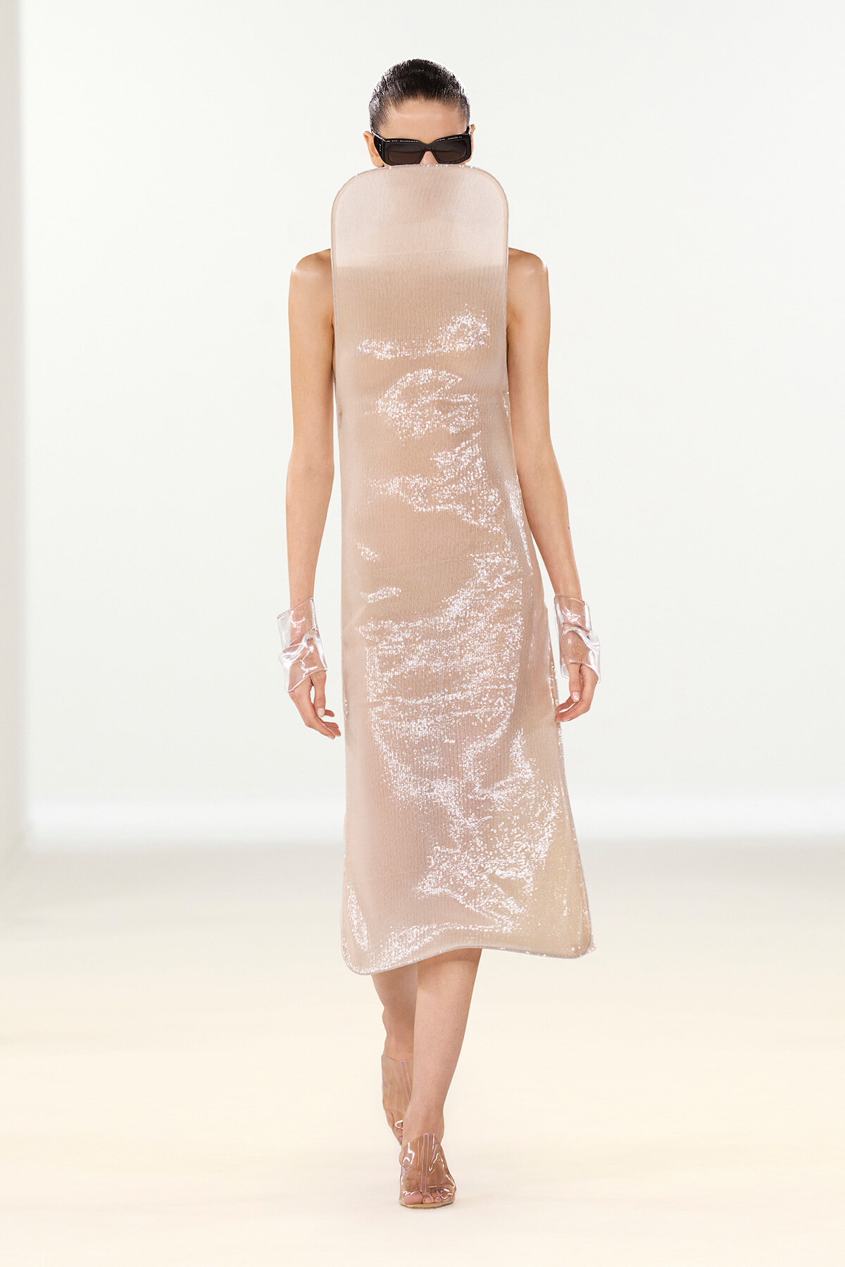 Runway model in a pale beige sequined midi dress with clear gloves and black sunglasses.