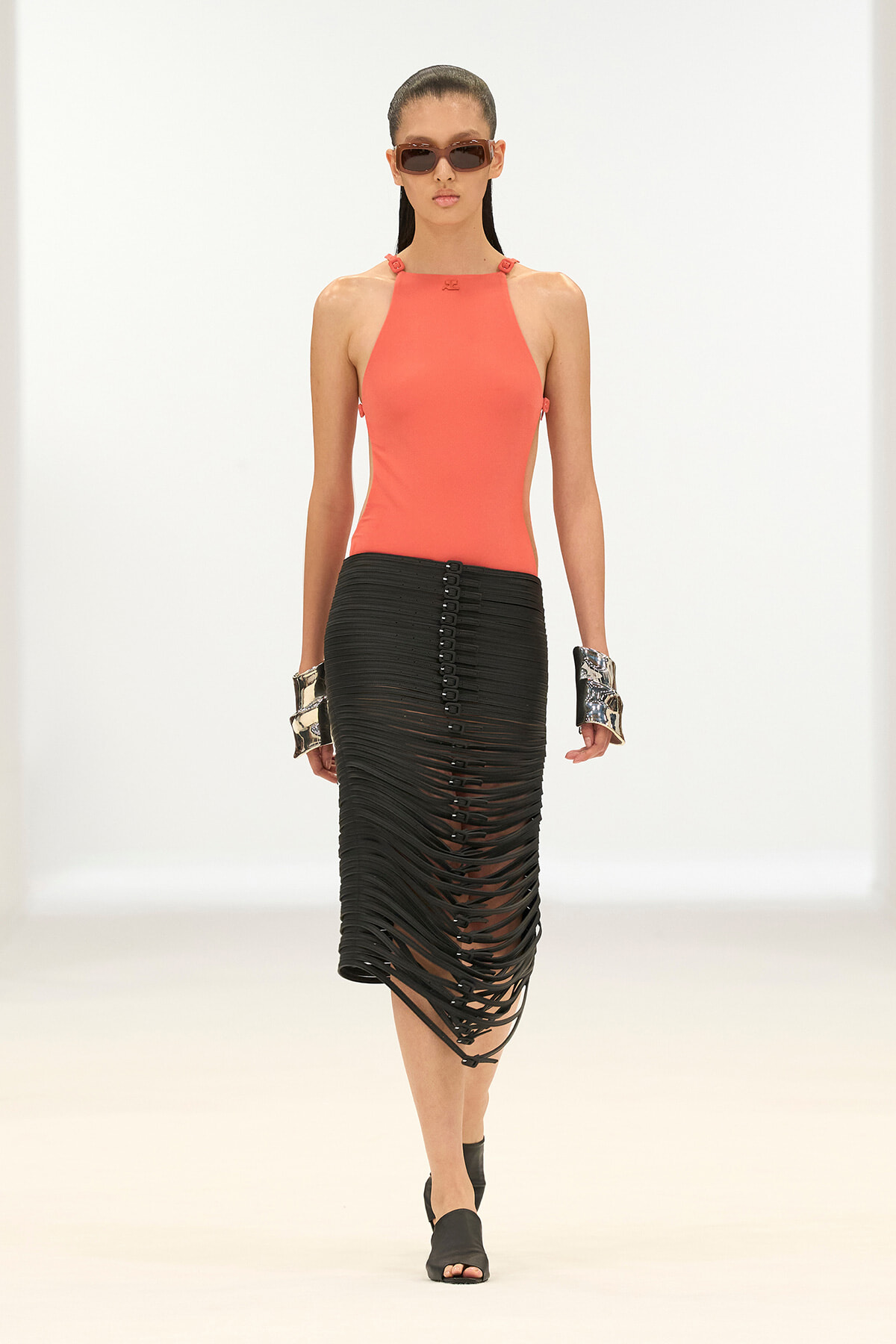 Model walking a runway in a coral halter top, black shredded midi skirt, sunglasses, and metallic cuffs on her wrists.