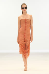 Model wearing a strapless orange dress with circular cutouts and matching sandals, walking on a white runway wearing black sunglasses.