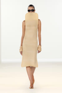Model on a runway wearing a beige midi dress with an oversized padded headpiece covering the face, plus sunglasses and matching cuffs on the wrists.
