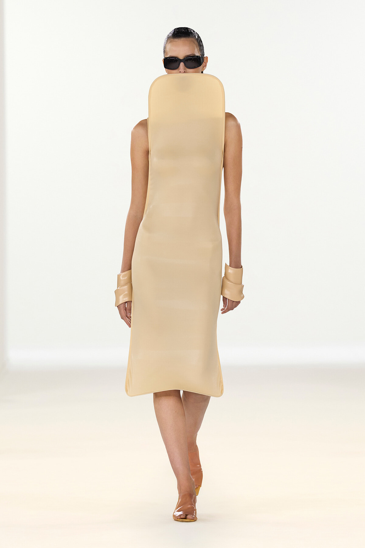 Model on a runway wearing a beige midi dress with an oversized padded headpiece covering the face, plus sunglasses and matching cuffs on the wrists.