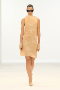 Model walks the runway in a sleeveless, peach sequin shift dress with large beige cuffs, nude boots, and black sunglasses.