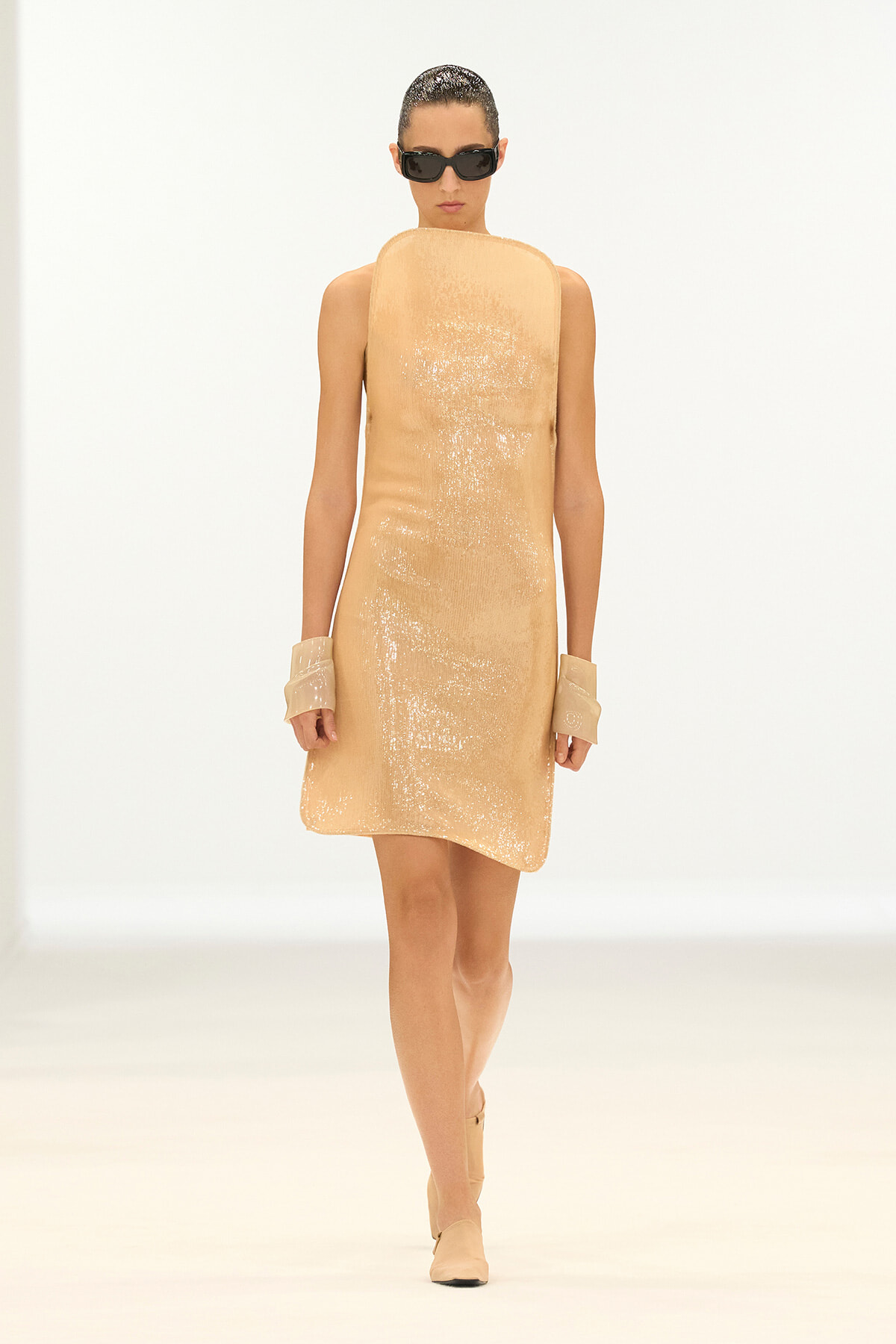 Model walks the runway in a sleeveless, peach sequin shift dress with large beige cuffs, nude boots, and black sunglasses.