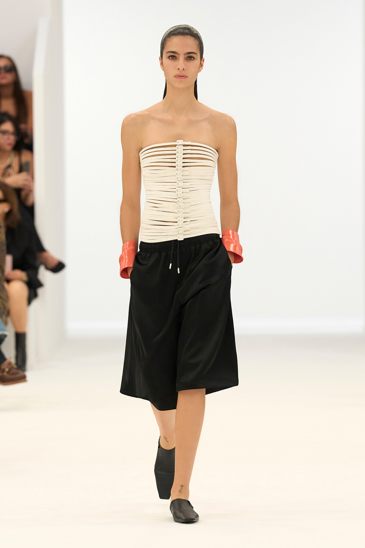 Model walking the runway in a strapless beige corset top with horizontal bands and black knee-length shorts, pink wrist cuffs, and black shoes.