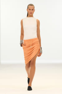 Model walking a white fashion runway in a sleeveless white top and peach ruched wrap skirt, wearing black flats and chunky silver bracelets.
