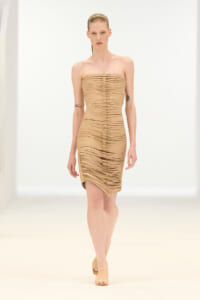 Model walking a runway in a nude-toned strapless, ruched midi dress with a vertical line of buttons down the center.
