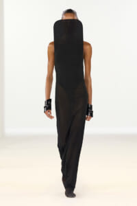 Back view of a model on a runway wearing an all-black ensemble with a mesh hood, sleeveless top, and sheer lower panel pants.