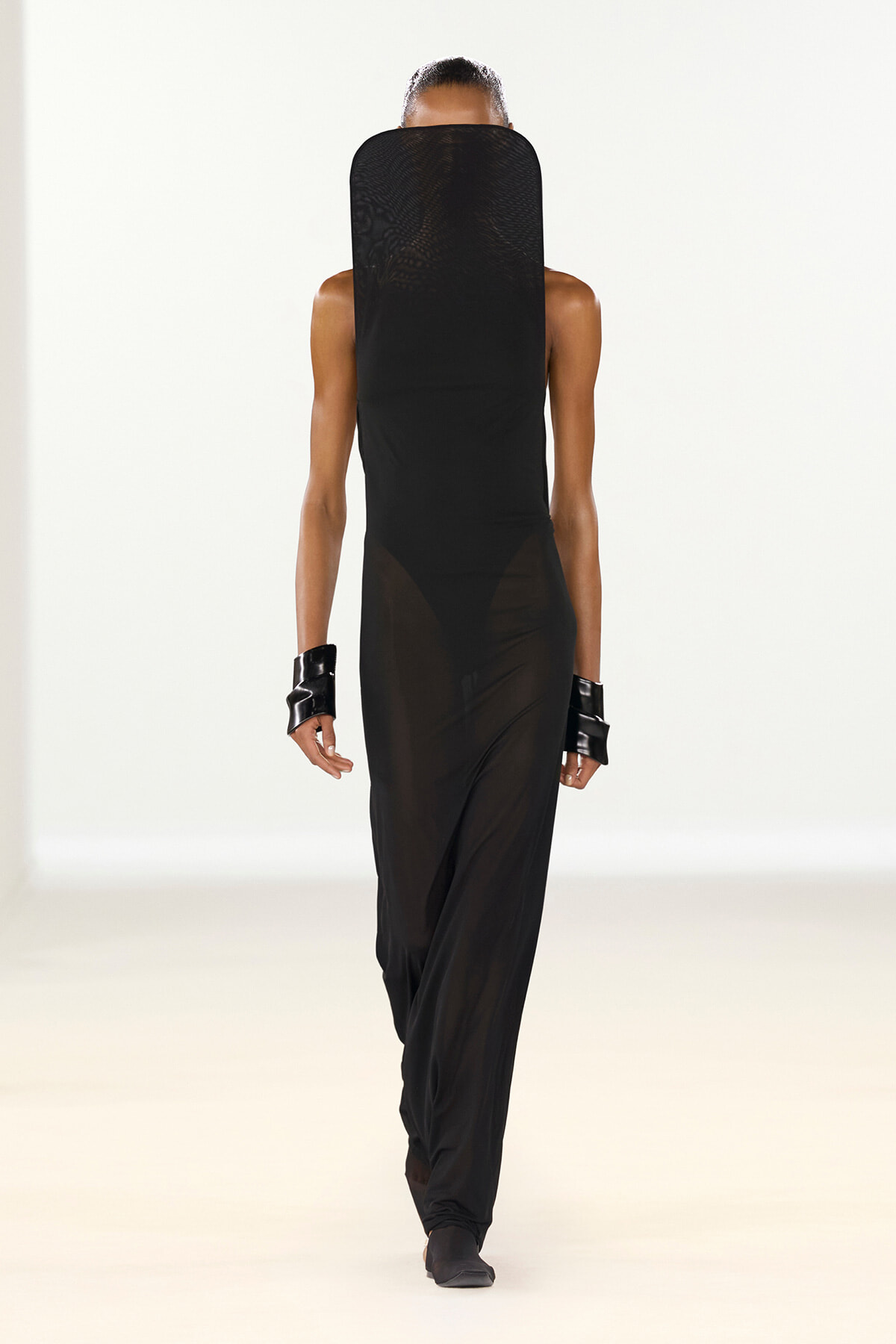 Back view of a model on a runway wearing an all-black ensemble with a mesh hood, sleeveless top, and sheer lower panel pants.