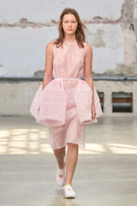 Female model walks a runway in a pale pink lace sleeveless dress with a voluminous sheer overlay skirt and matching shoes.