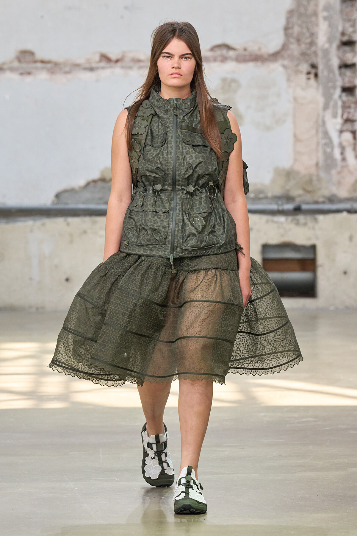 Model on a runway wearing an olive-green lace midi skirt with a matching utility vest and floral appliqués on the shoulders/placeholders.