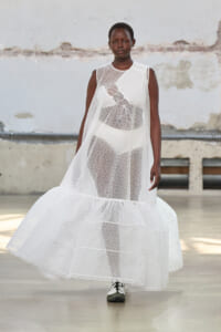 Model walking a runway in a long, sheer white lace dress with a tiered, voluminous hem against an industrial backdrop, full-length view.