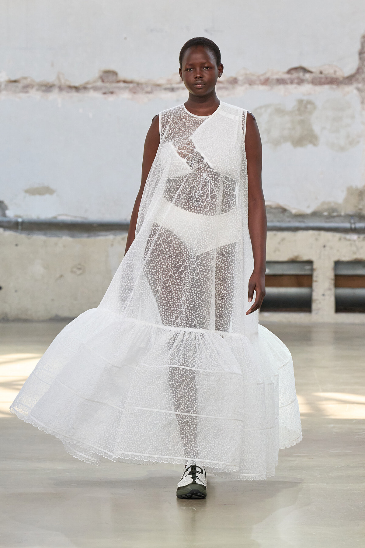 Model walking a runway in a long, sheer white lace dress with a tiered, voluminous hem against an industrial backdrop, full-length view.