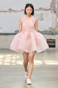 Model walking a runway in a pale pink lace dress with a voluminous tiered skirt and sheer bodice.