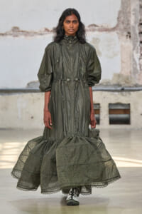 Model walking the runway in an olive-green, zip-front dress with a gathered waist and a voluminous lace-trimmed hem.