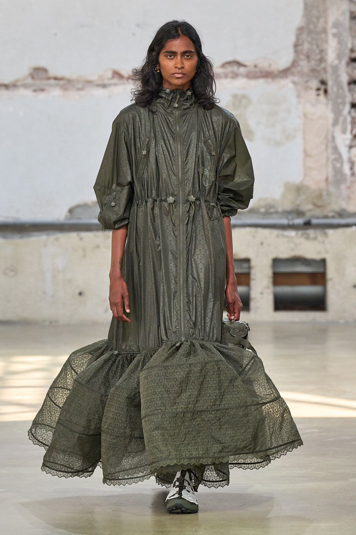 Model walking the runway in an olive-green, zip-front dress with a gathered waist and a voluminous lace-trimmed hem.