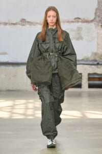 Model walking a runway in an olive-green camouflage oversized jacket and matching pants with a belt.
