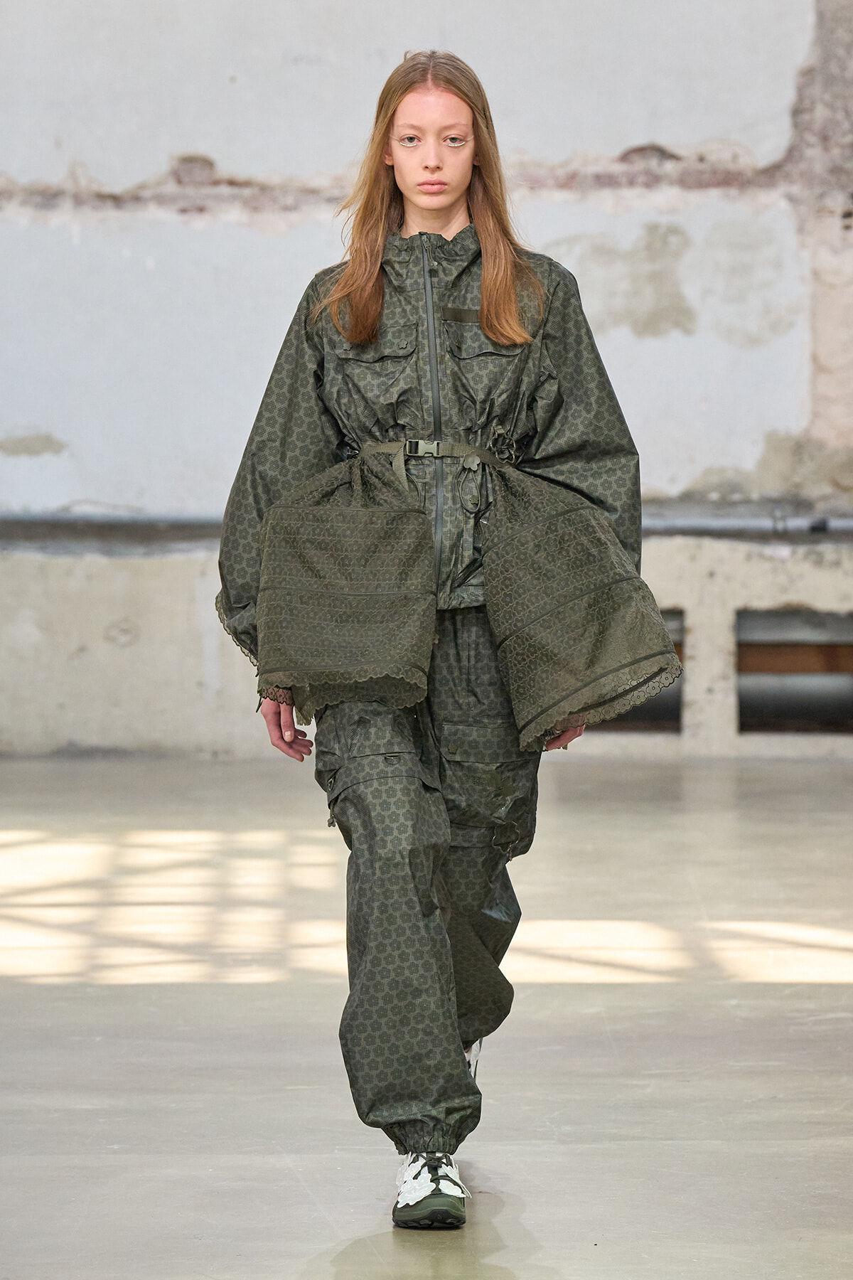 Model walking a runway in an olive-green camouflage oversized jacket and matching pants with a belt.