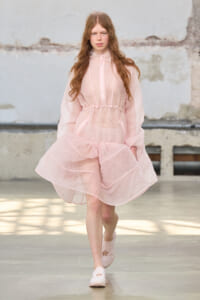 Model walking the runway in a light pink, sheer dress with a drawstring waist and long sleeves, in a minimalist setting.