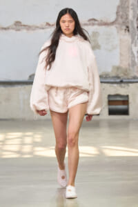 Model in oversized pale pink hoodie and satin shorts walking a fashion show runway.