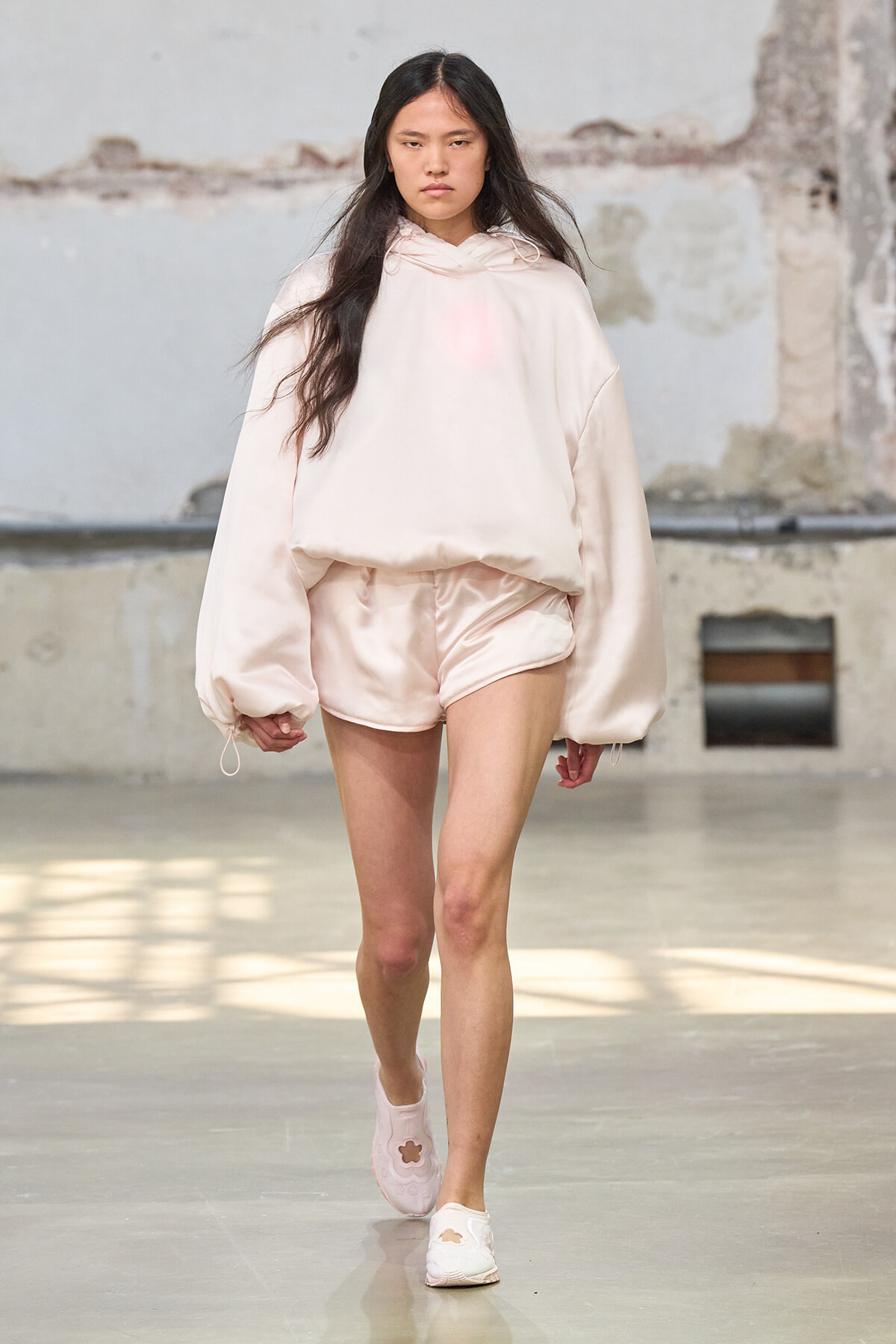 Model in oversized pale pink hoodie and satin shorts walking a fashion show runway.