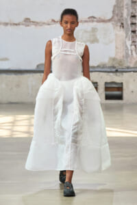 Fashion model wearing a voluminous white textured dress with sheer bodice, walking toward the camera in an industrial space.
