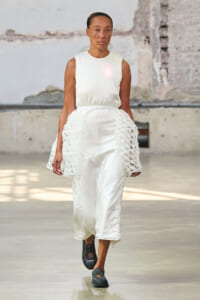 Model walking a runway in an all-white outfit: sleeveless top and satin wide-leg pants with crochet side panels, in an industrial venue.
