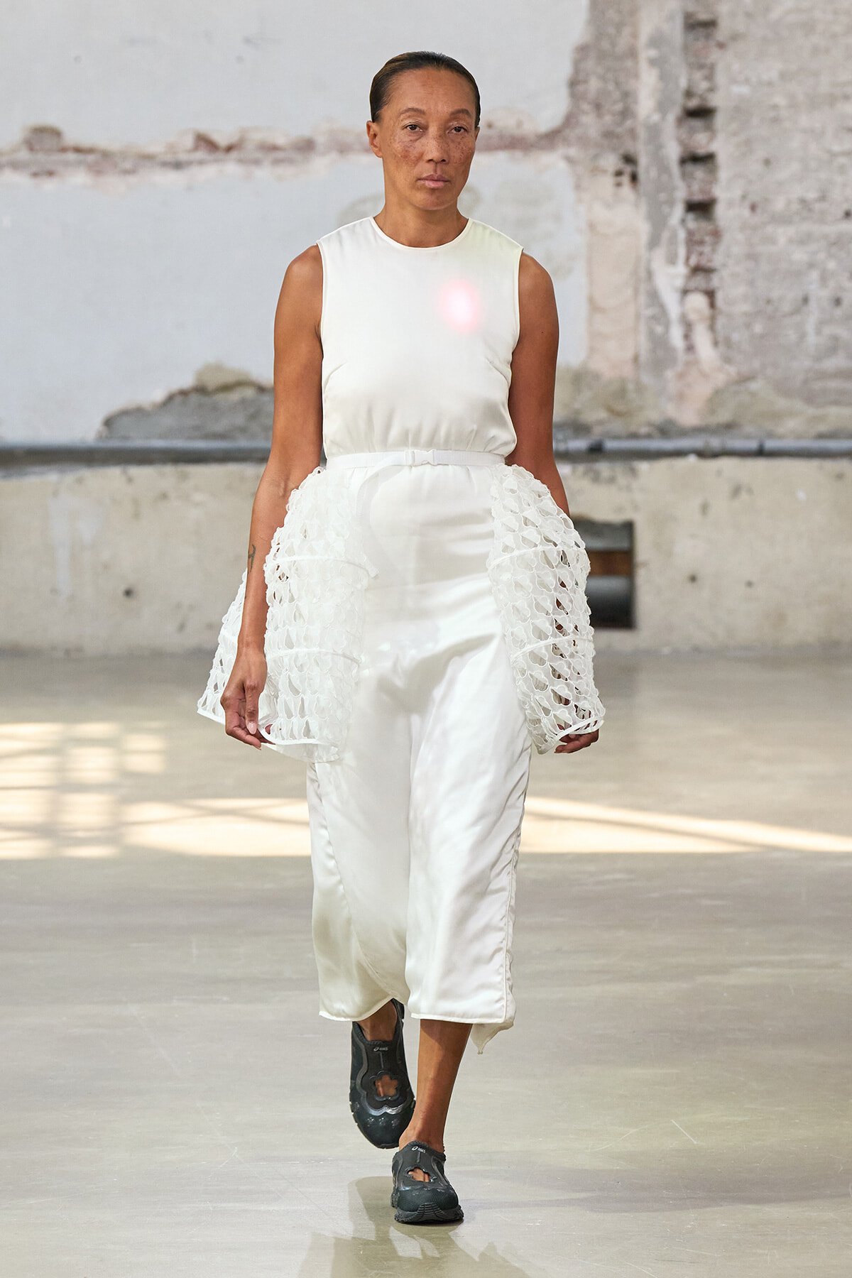Model walking a runway in an all-white outfit: sleeveless top and satin wide-leg pants with crochet side panels, in an industrial venue.