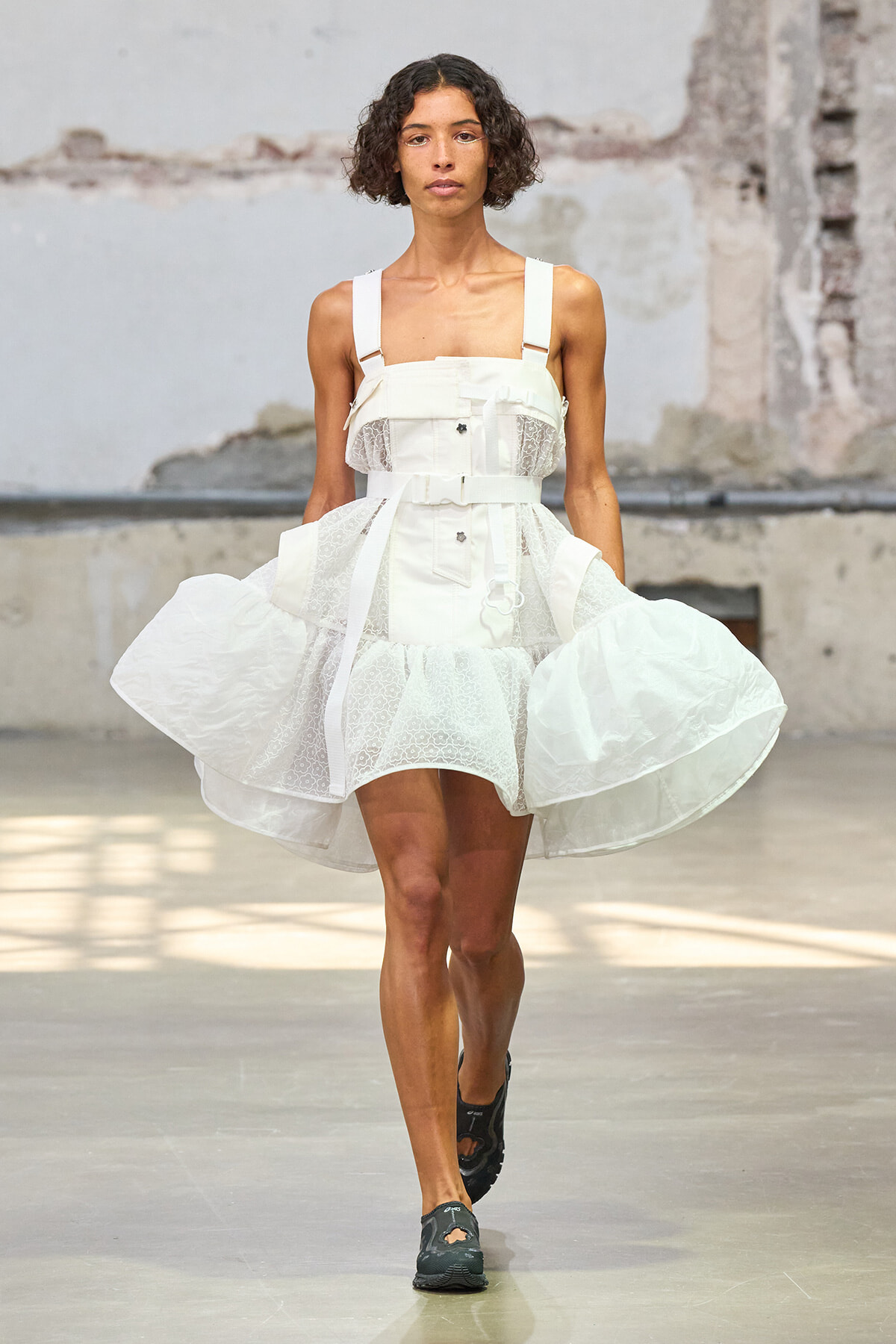 Model on a runway wearing a white structured dress with lace panels and a thick waist belt in an industrial venue.