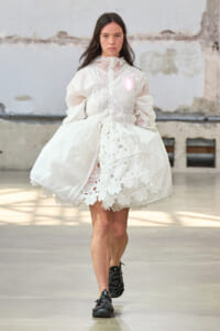 Model walking a runway in an oversized white dress with a sheer high neck, corset lacing, and a floral lace hem, paired with black ankle boots.