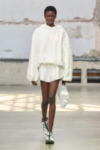 Model walks a runway in an oversized white hoodie and matching shorts, holding a white bag at her side.