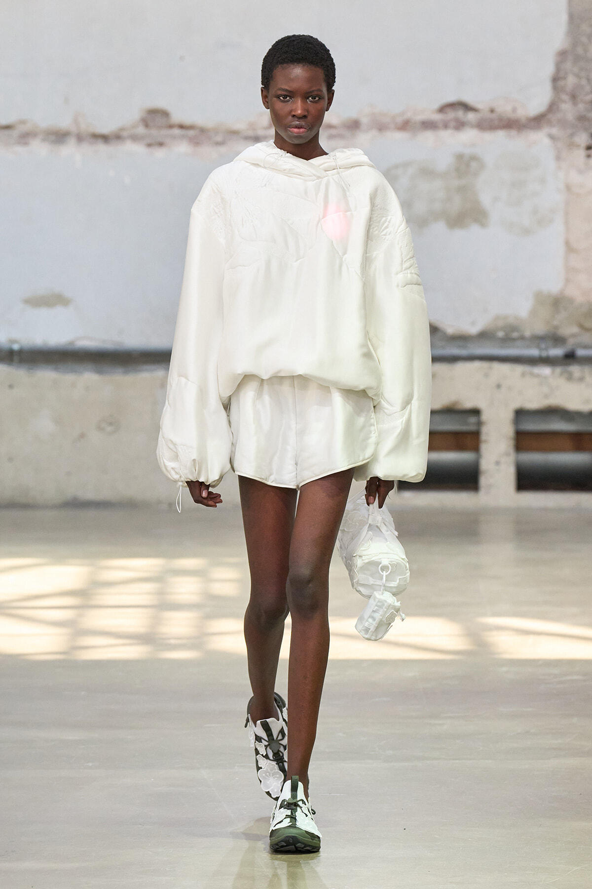 Model walks a runway in an oversized white hoodie and matching shorts, holding a white bag at her side.