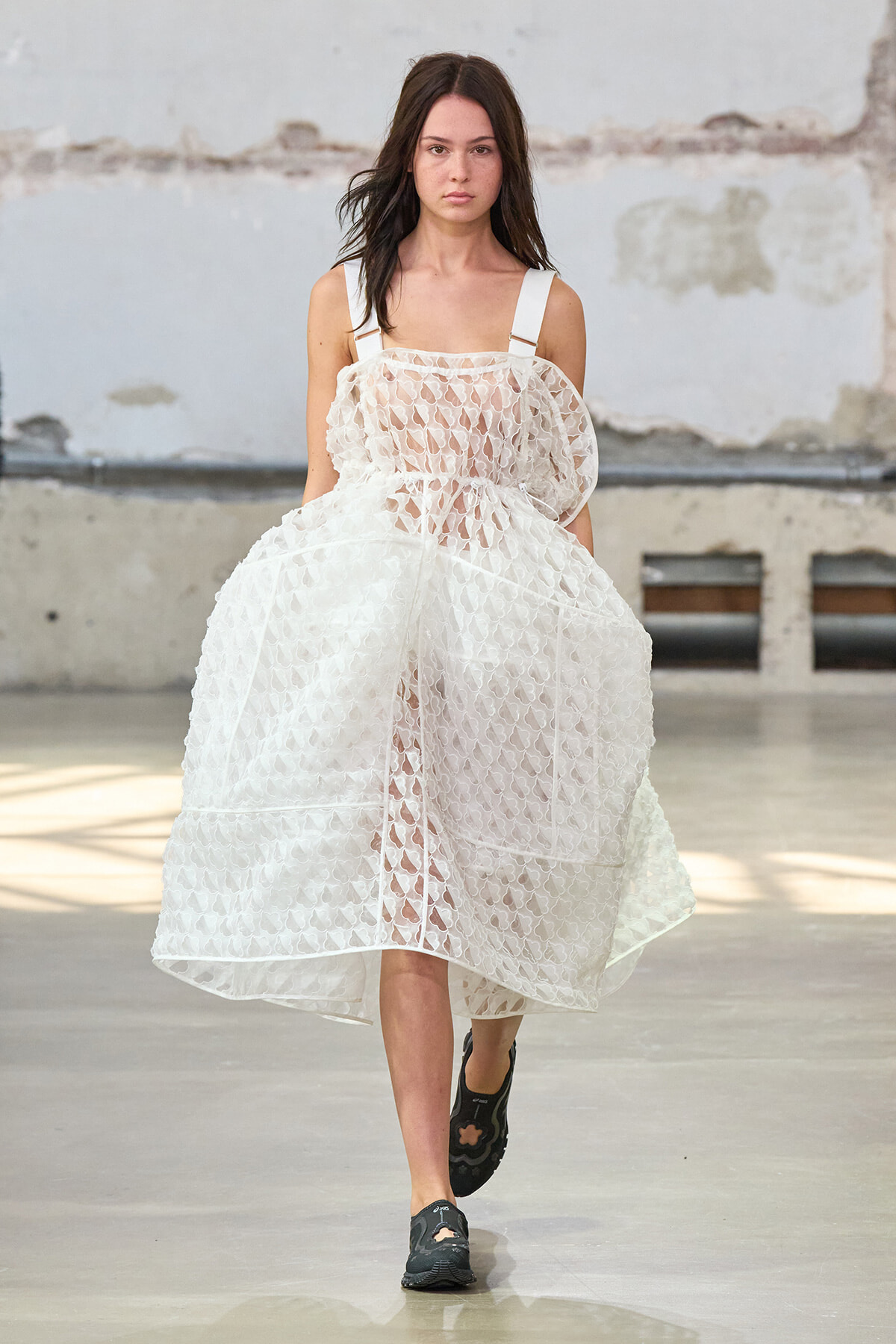 Model walking a runway in a white, lace tiered dress with broad shoulder straps and black sneakers in an industrial-styled studio.