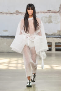 Model walking a fashion show in a sheer white lace poncho and matching pants, carrying a white bag with floral details.