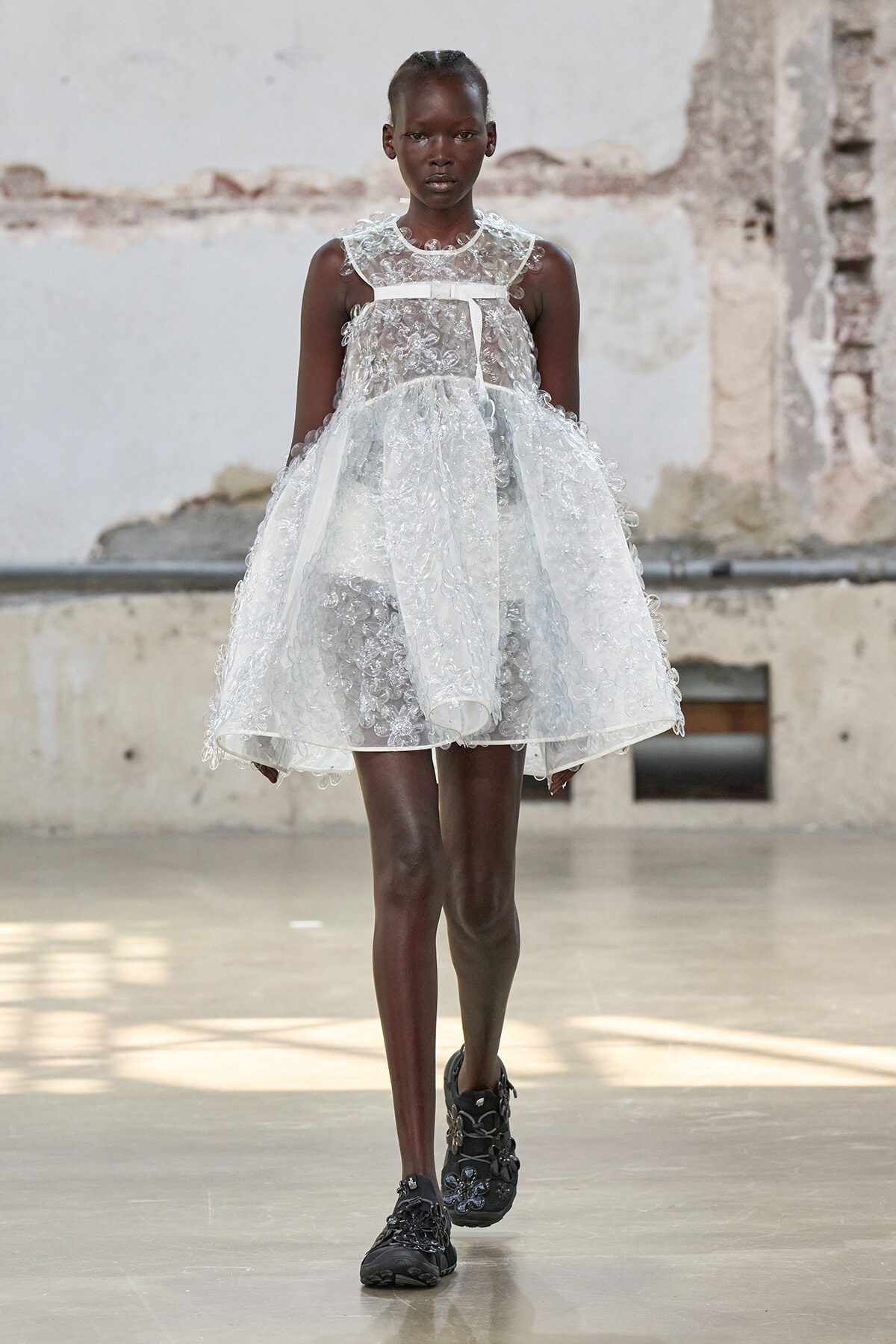 Model in a white lace, knee‑length dress with a sheer overlay and boots on a concrete runway backdrop.