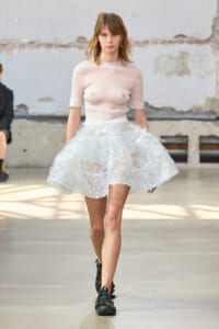 Model walking the runway in a sheer pale-pink top and a voluminous white lace bubble skirt, paired with black boots in an industrial setting.