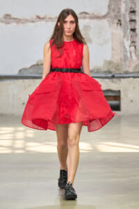 Runway model wearing a bright red sleeveless dress with a black belt, walking in an industrial-style venue.