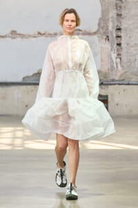 Woman wearing a sheer white hooded dress with a cinched waist and voluminous skirt, walking in an industrial space.