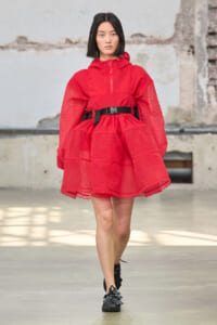 Model wearing a red lace-overlay dress with hood and black waist belt, walking on a runway in an industrial venue.